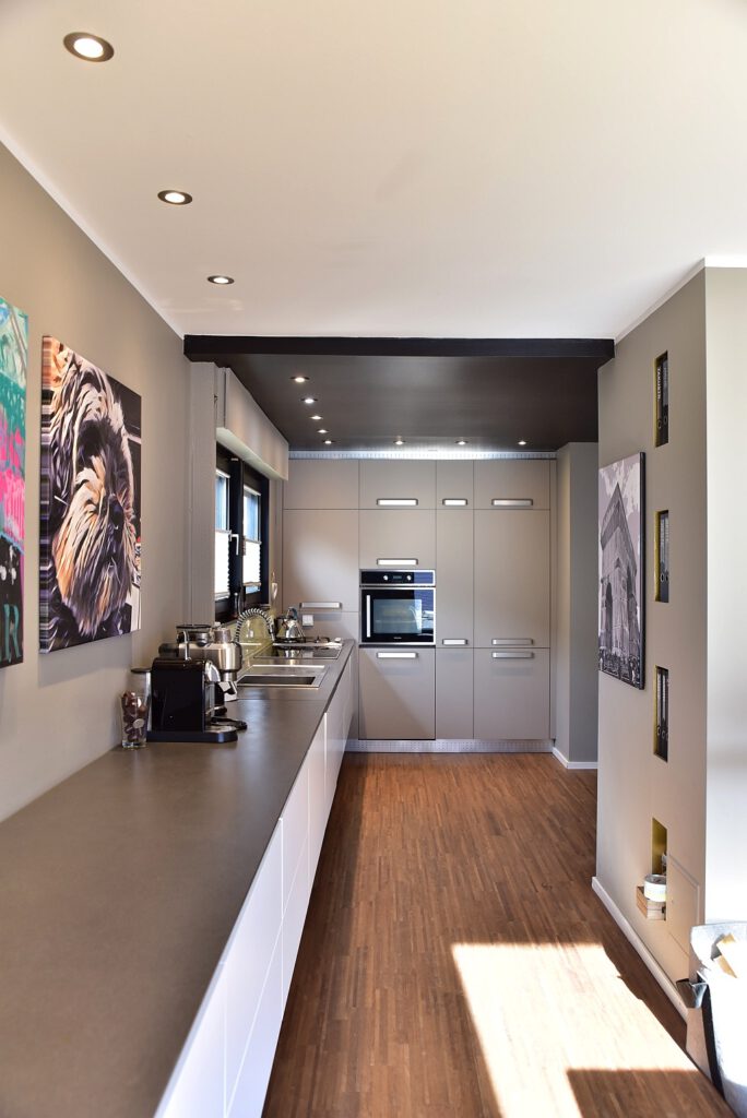 Lange, moderne Küchenzeile in Grau mit grifflosen Fronten und einer dunklen Deckeninstallation im minimalistischen Stil.