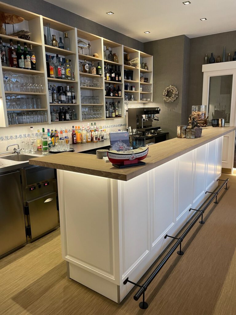 Schräge Ansicht einer maßgefertigten weißen Bartheke mit Massivholz-Abdeckung und beleuchtetem Rückbuffet-Regal von Schreinerei Pass-genau.