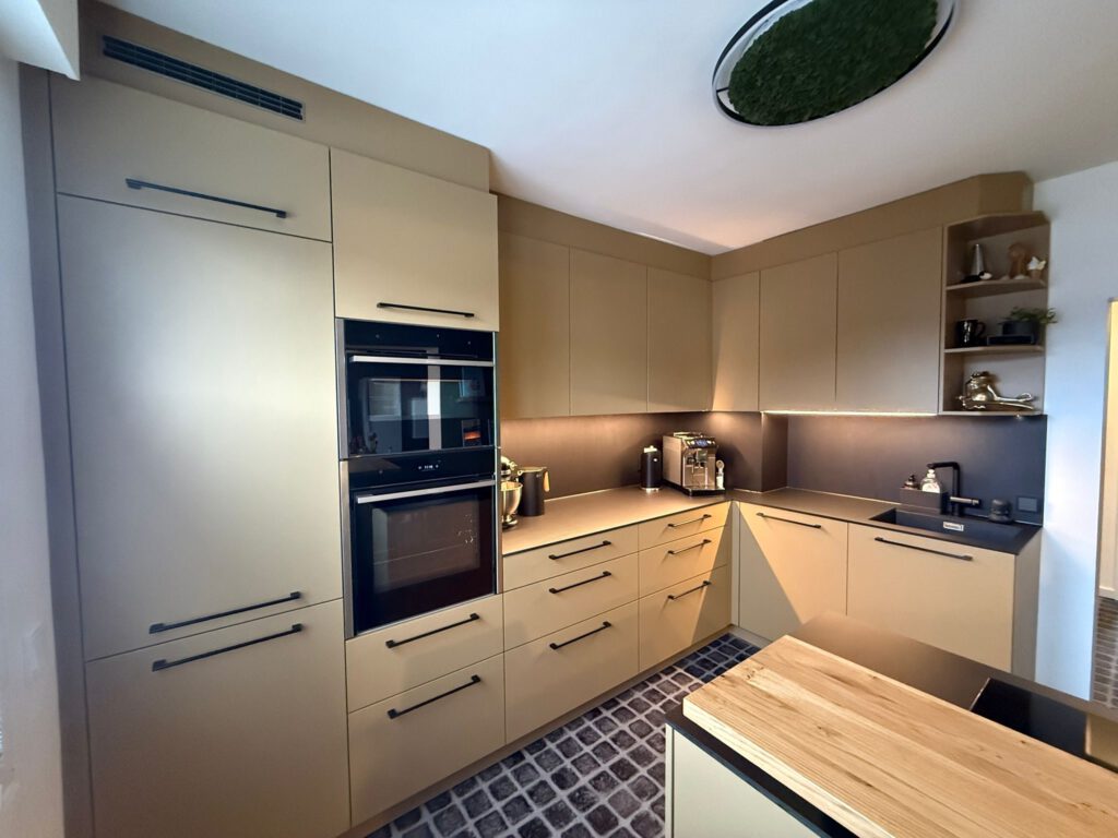 Weite Ansicht einer sandfarbenen Design-Küche mit Kochinsel und einem integrierten Holzelement als Arbeitsplatte